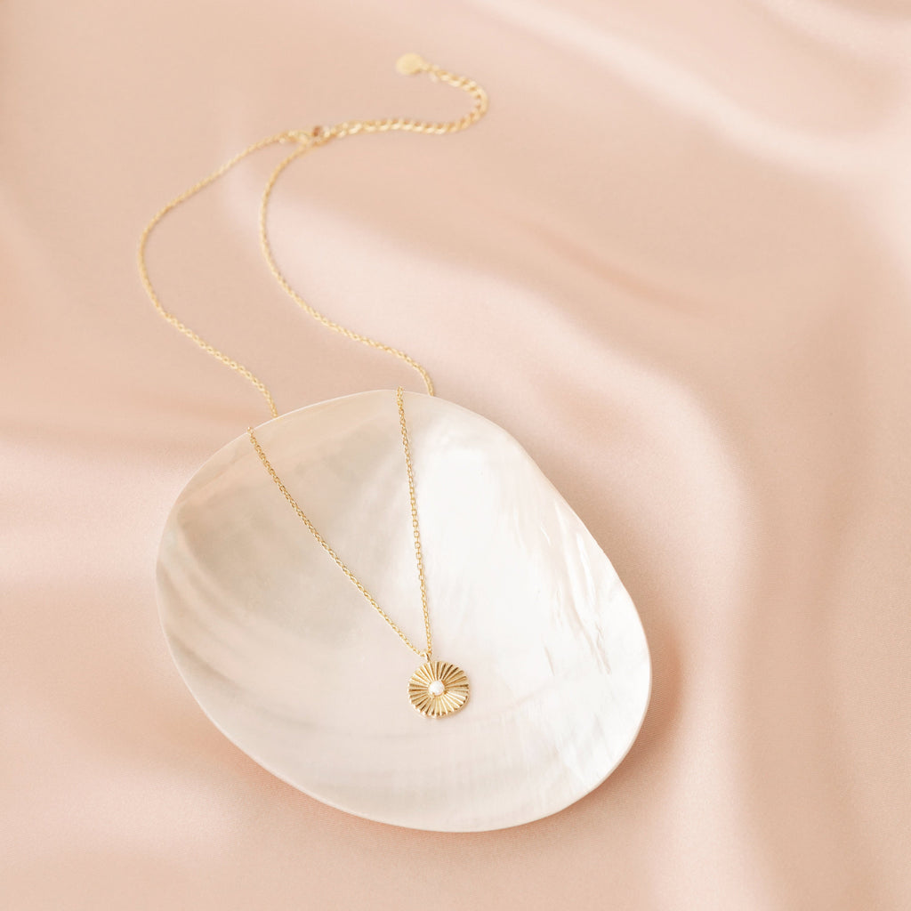 Gold opal medallion necklace with a delicate chain and a sunburst pendant featuring a luminous opal center, styled on a white shell dish atop a soft blush satin background. 