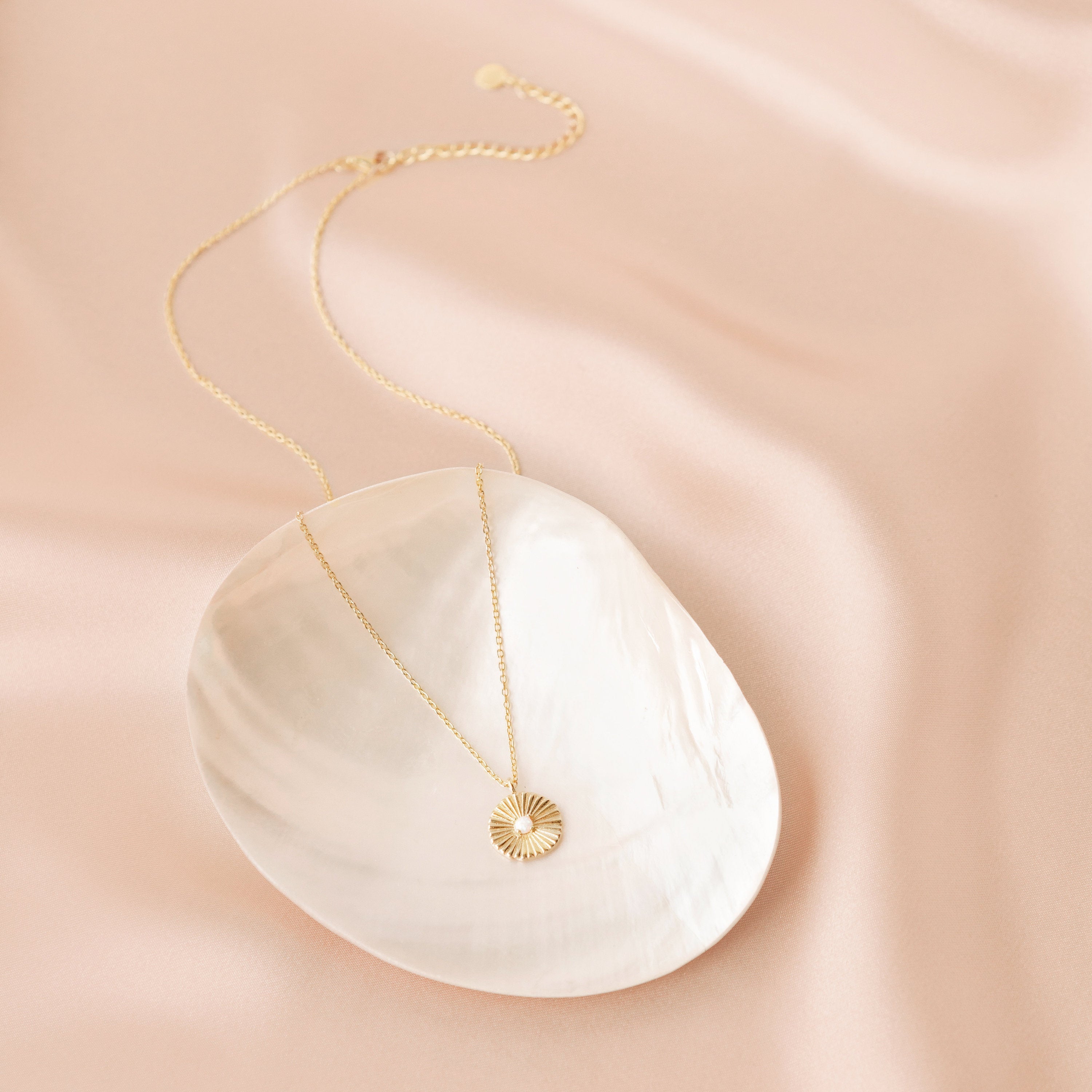 Gold opal medallion necklace with a delicate chain and a sunburst pendant featuring a luminous opal center, styled on a white shell dish atop a soft blush satin background. 