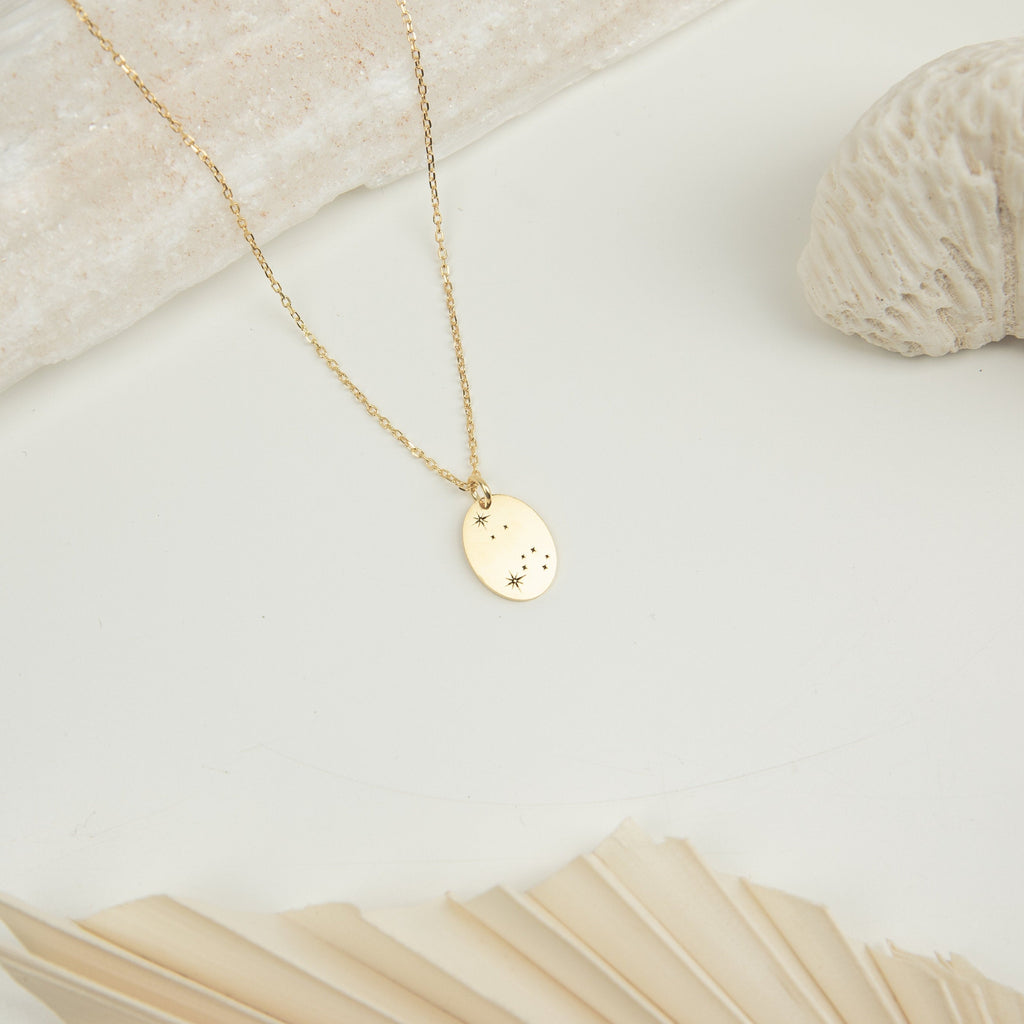 Zodiac Constellation Necklace: a gold oval pendant with star engravings, shown on a minimal light background with shells.