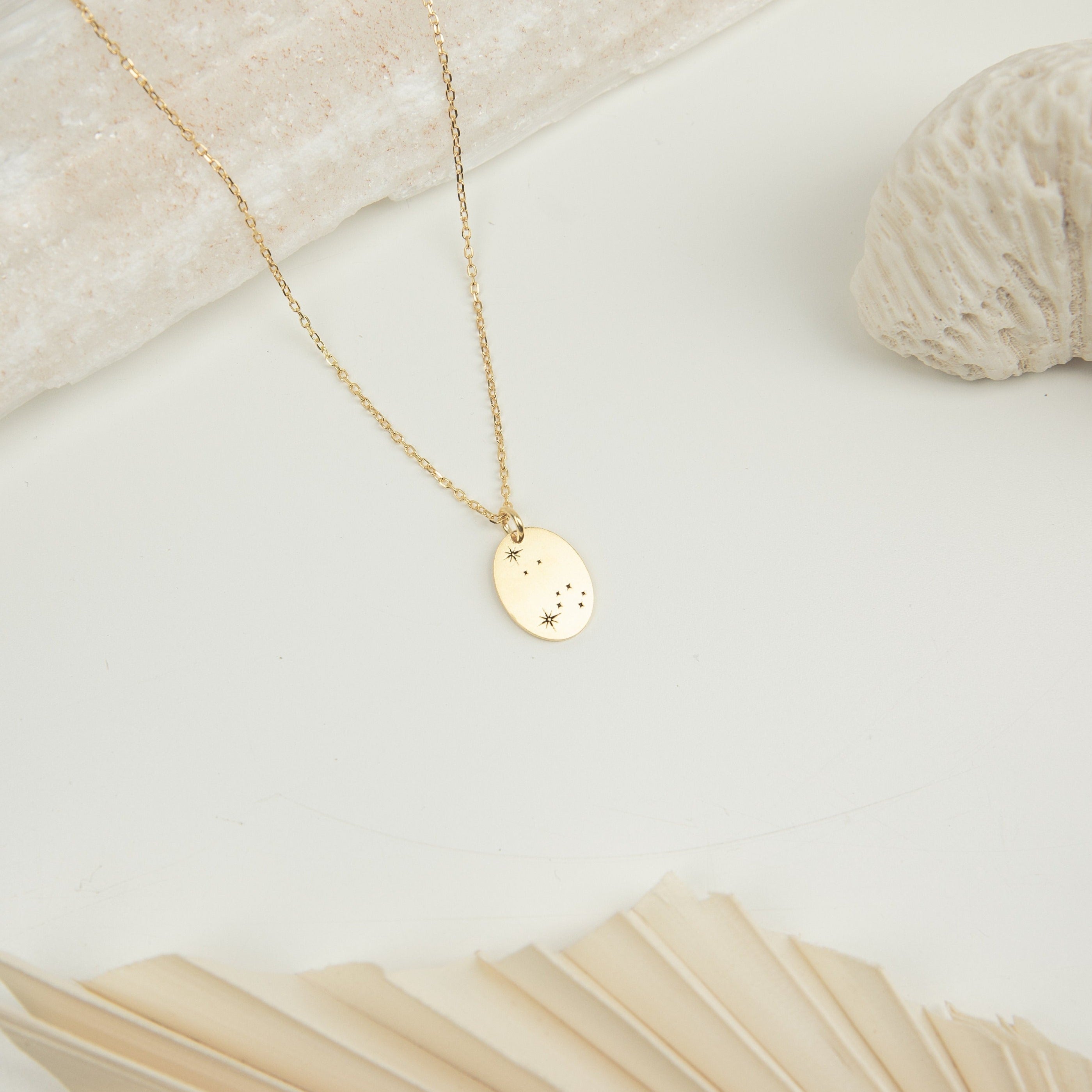 Zodiac Constellation Necklace: a gold oval pendant with star engravings, shown on a minimal light background with shells.