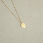 The Zodiac Constellation Necklace features an oval gold pendant engraved with small star and dot designs, displayed on a light beige background.