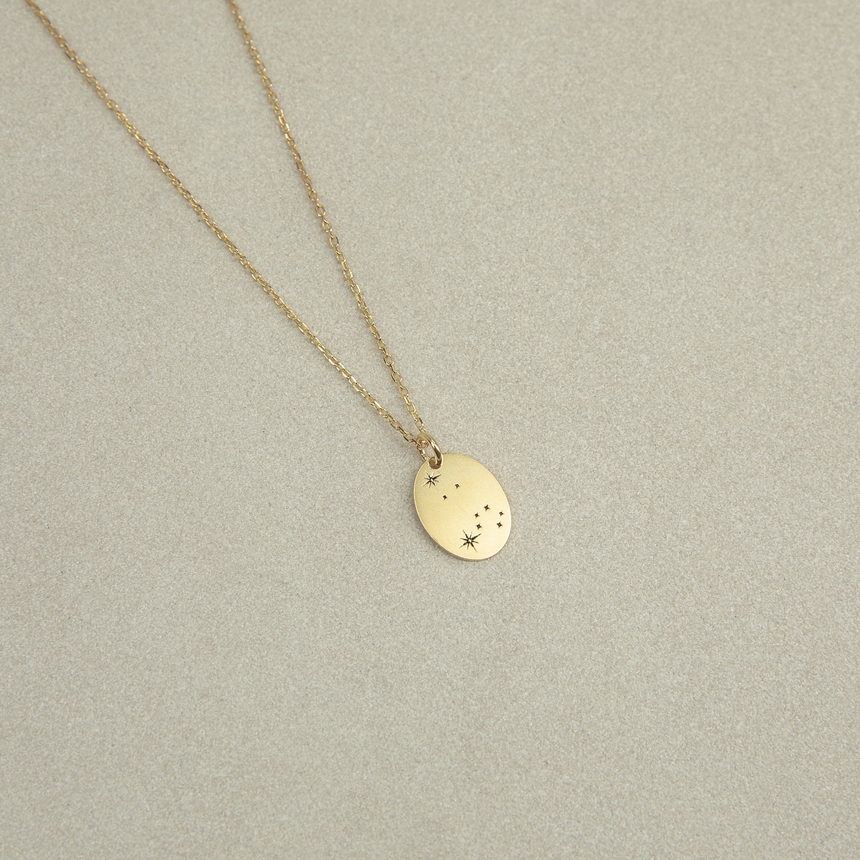 The Zodiac Constellation Necklace features an oval gold pendant engraved with small star and dot designs, displayed on a light beige background.