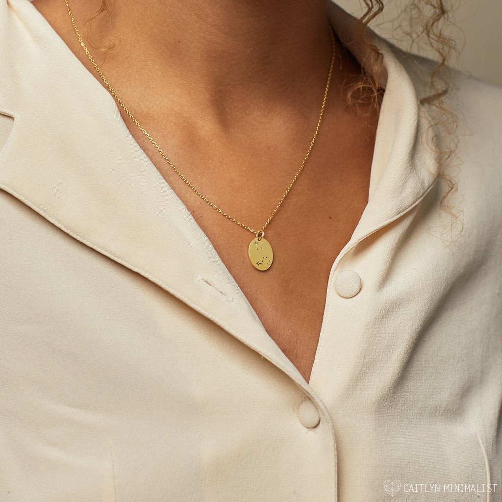 A woman wears a cream blouse and the Zodiac Constellation Necklace featuring an oval pendant with engraved initials.
