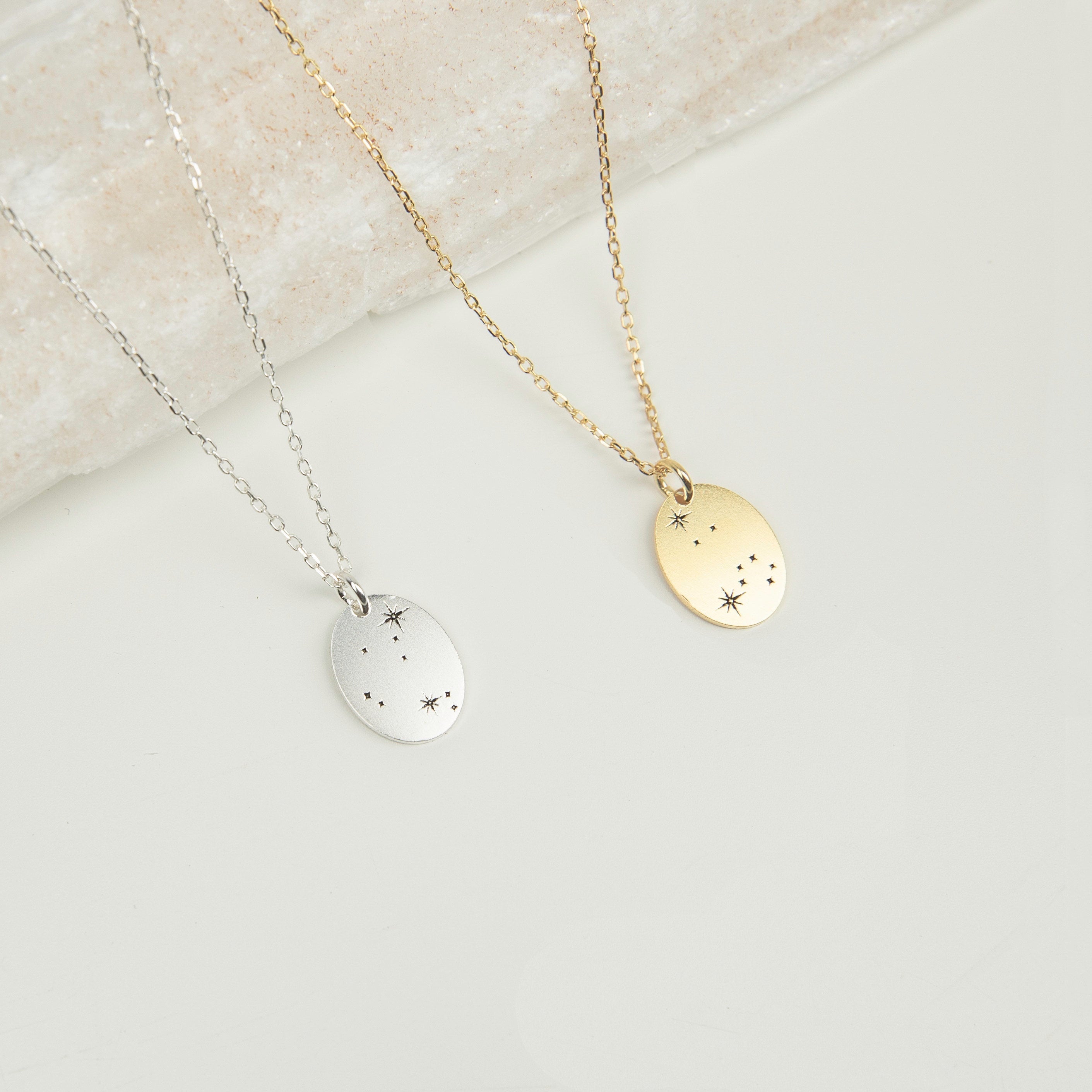 The Zodiac Constellation Necklace features oval pendants in silver and gold, each engraved with delicate star and dot designs, capturing the allure of constellation-inspired amulets.