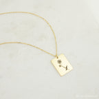 The Birth Flower Tag Necklace features a rectangular gold pendant engraved with a rose, displayed on a light textured surface—an ideal choice for personalized birth month jewelry.