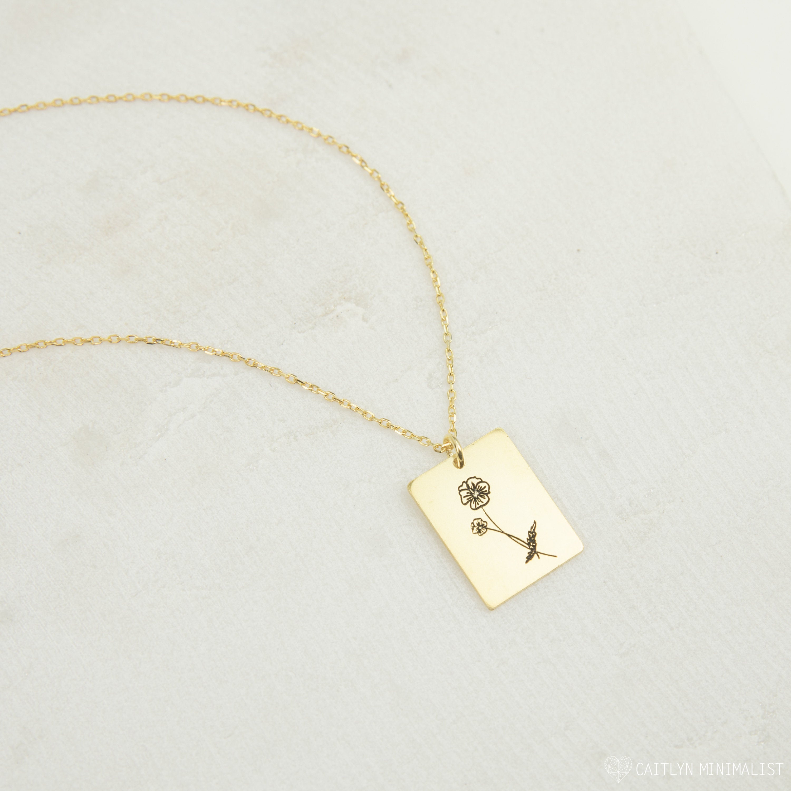The Birth Flower Tag Necklace features a rectangular gold pendant engraved with a rose, displayed on a light textured surface—an ideal choice for personalized birth month jewelry.
