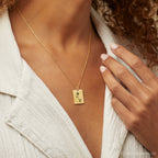 A woman wears the Birth Flower Tag Necklace, featuring a gold rectangular pendant engraved with a rose design, styled with a light jacket.