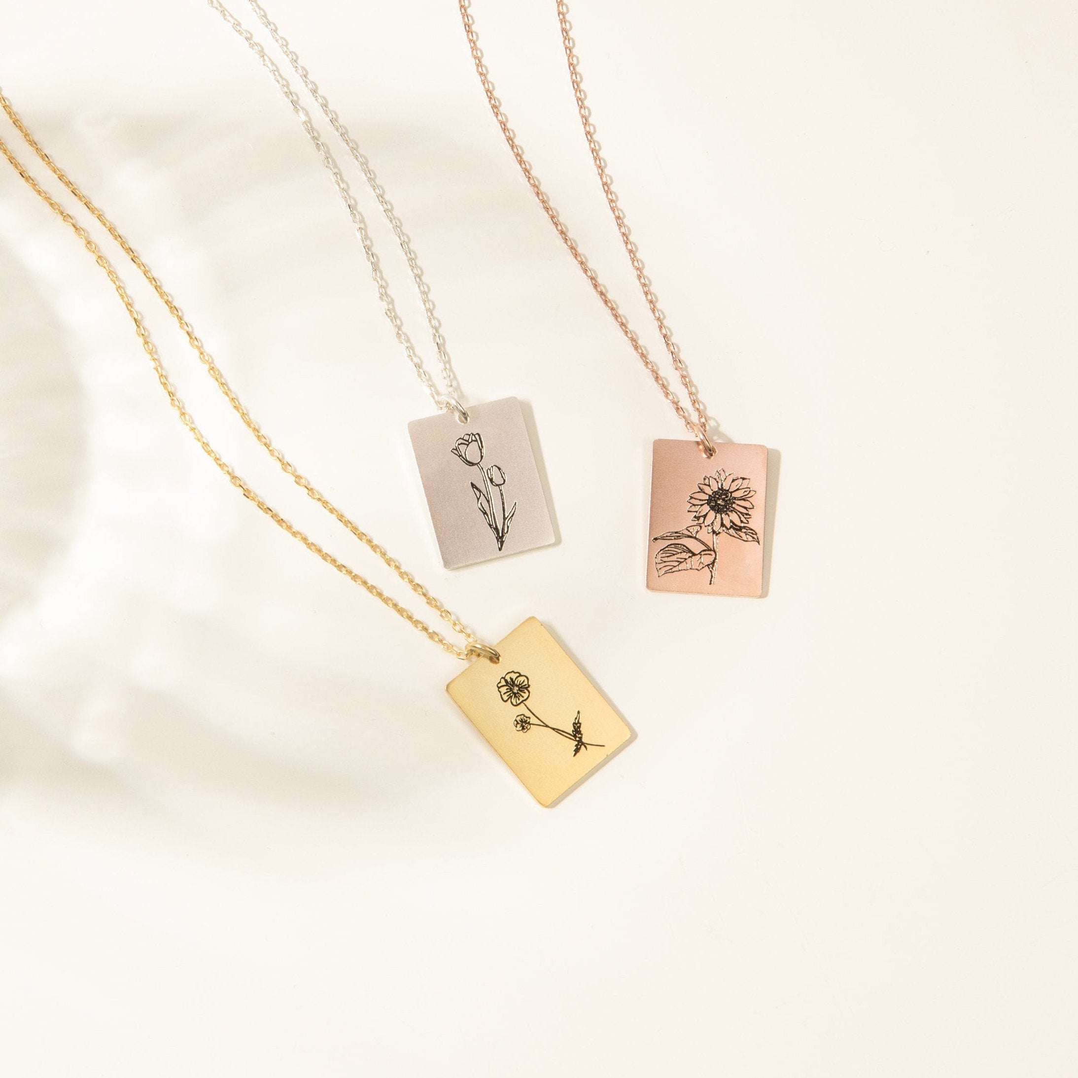 The Birth Flower Tag Necklace comes in gold, silver, and rose gold with engraved floral designs—ideal as personalized jewelry or a meaningful birth month gift. Shown are three necklaces on a white background.