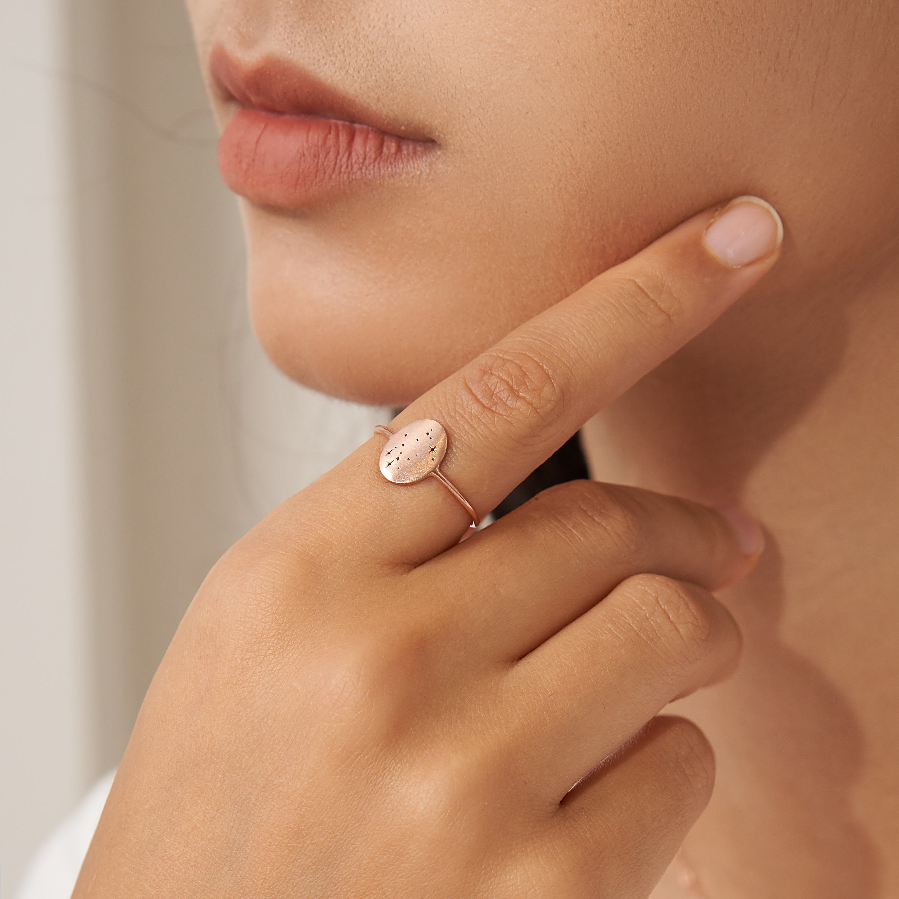 A person touches their face while wearing the Constellation Oval Ring, a gold piece featuring an oval-shaped, textured design on their finger.