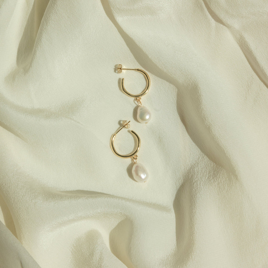 The Margot Pearl Hoops, featuring elegant drop pearls on gold hoops, rest on soft, light cream-colored fabric.