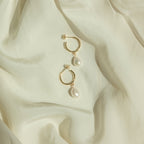 The Margot Pearl Hoops, featuring elegant drop pearls on gold hoops, rest on soft, light cream-colored fabric.