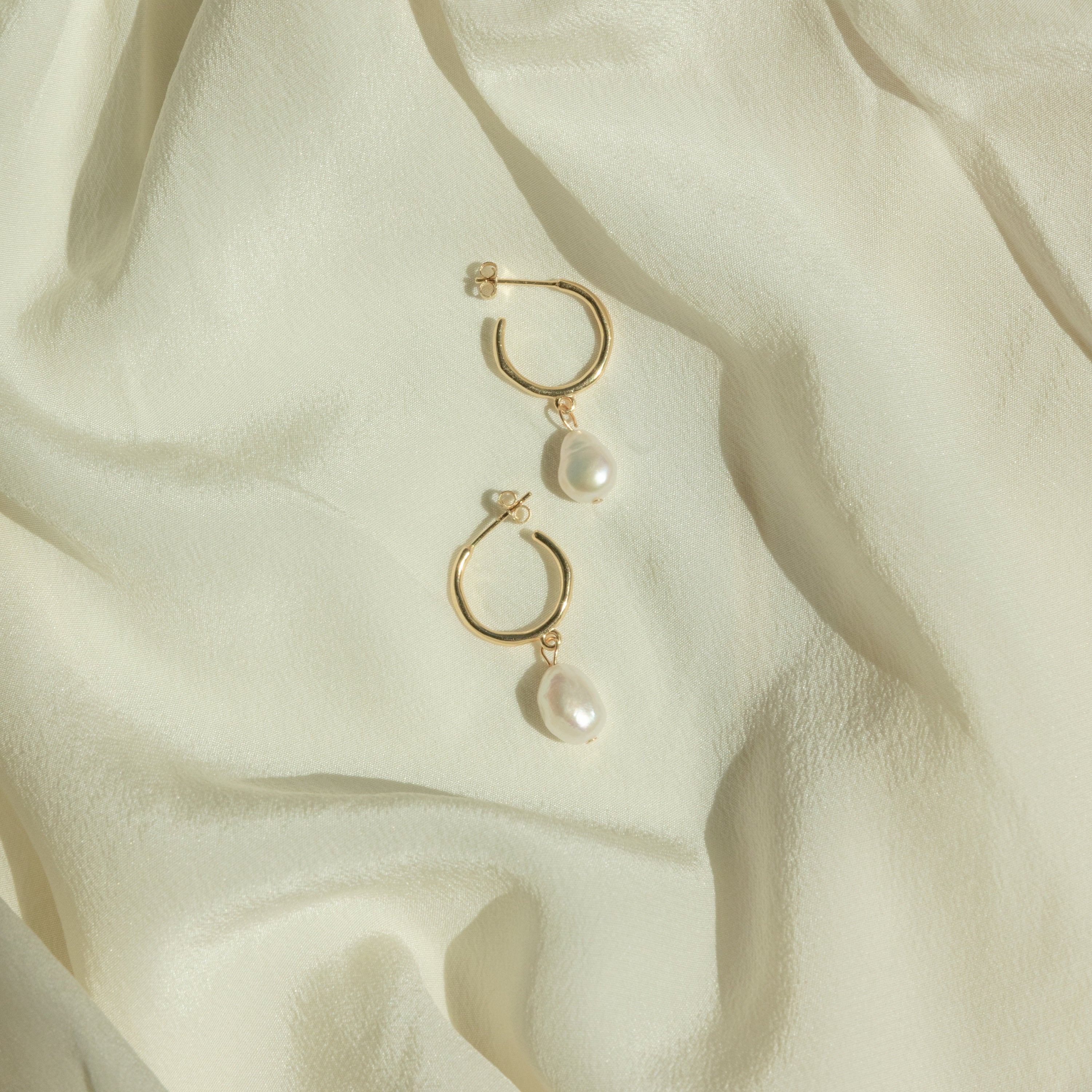 The Margot Pearl Hoops, featuring elegant drop pearls on gold hoops, rest on soft, light cream-colored fabric.