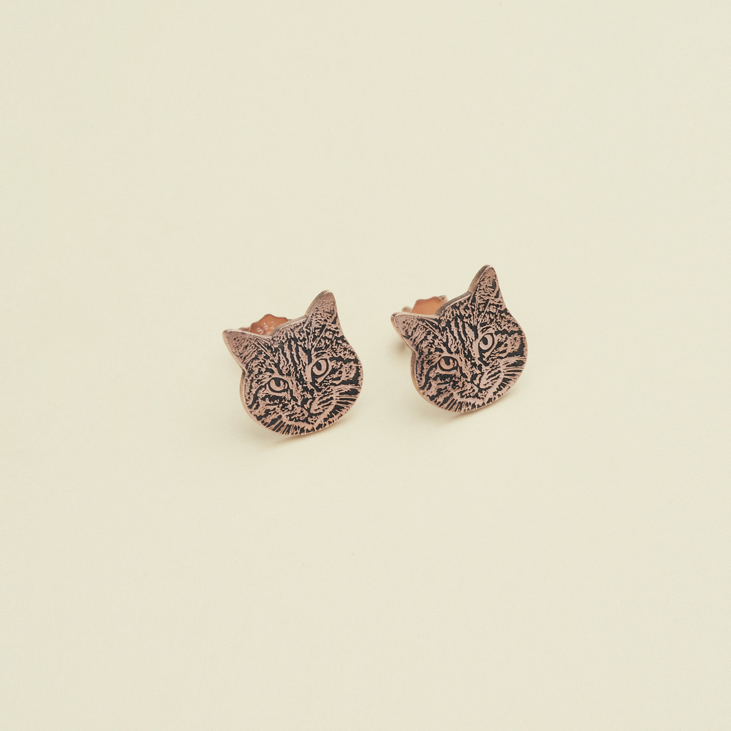 The Pet Portrait Studs, a pair of copper cat face earrings ideal for pet lovers, are showcased on a plain cream background.