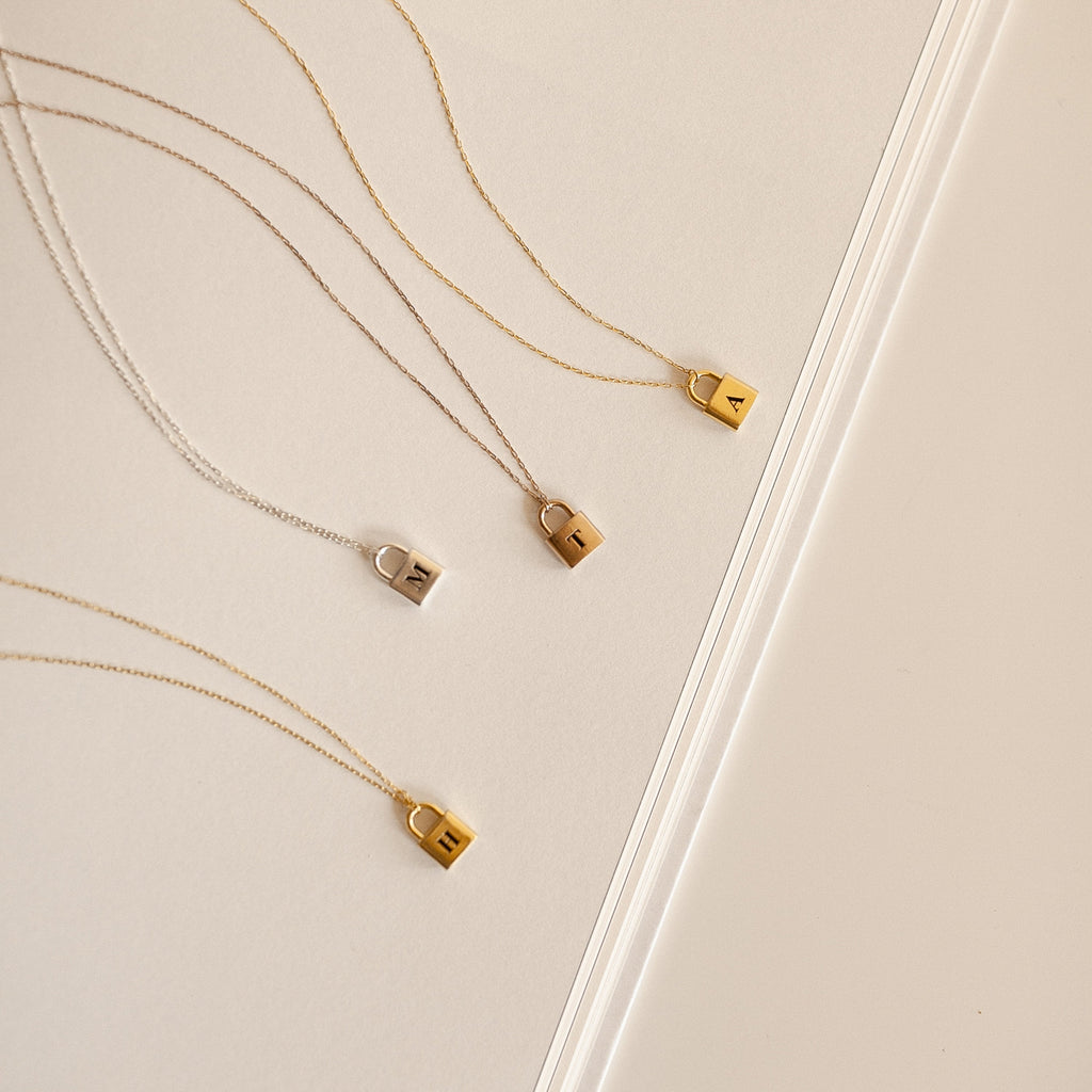 The Padlock Necklace in Rose Gold features a small lock pendant engraved with an initial, displayed on a light background—ideal for a subtle, personalized fashion statement.