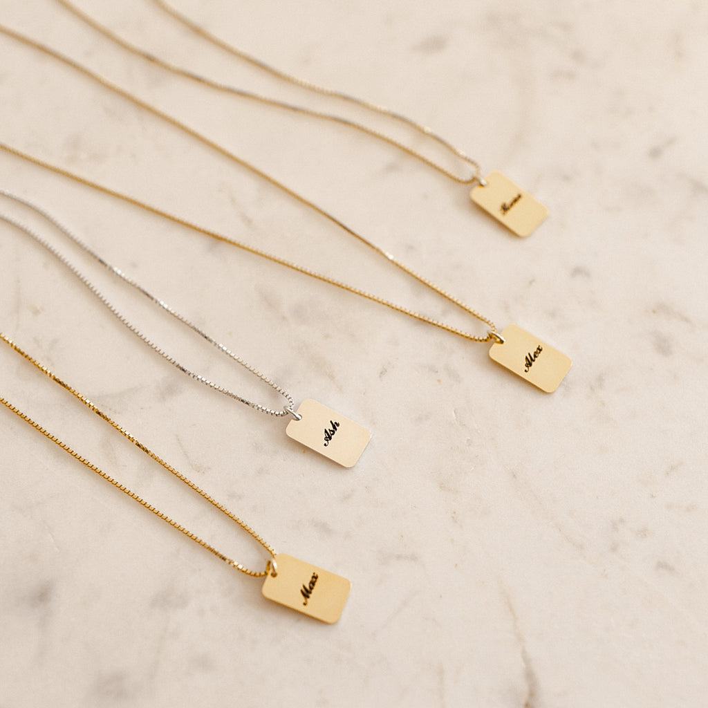 Four rectangular Name Tag Necklaces in gold and silver with engraved names are displayed on a light marble surface, highlighting personalized name jewelry.