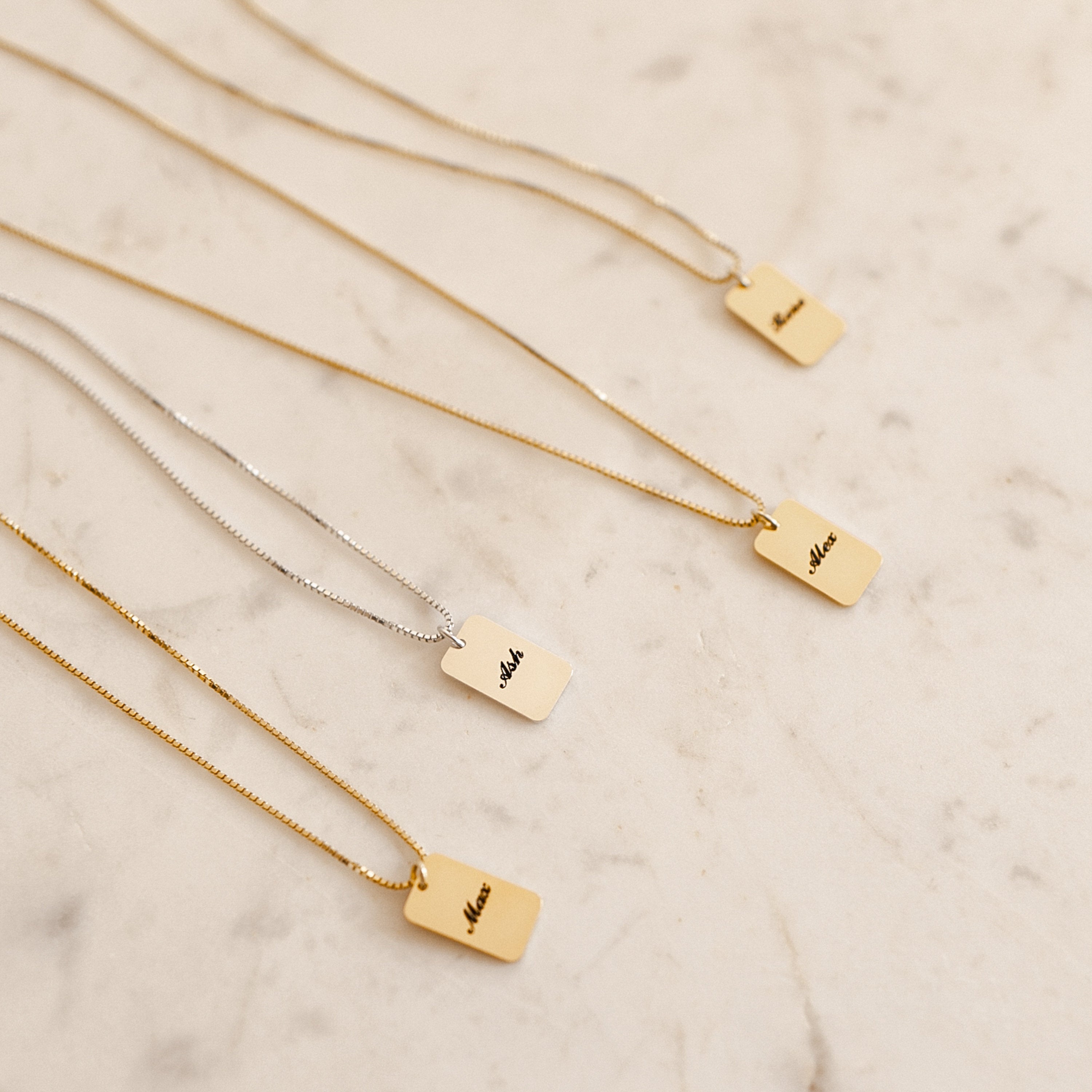 Four rectangular Name Tag Necklaces in gold and silver with engraved names are displayed on a light marble surface, highlighting personalized name jewelry.