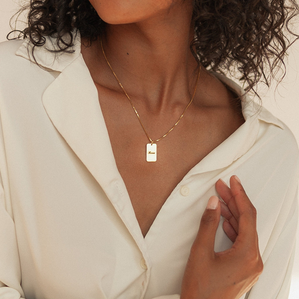 A woman in a cream blouse wears the Name Tag Necklace in Rose Gold, featuring a rectangular pendant, with her hand lifted near her collar.