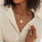 A woman in a cream blouse wears the Name Tag Necklace in Rose Gold, featuring a rectangular pendant, with her hand lifted near her collar.