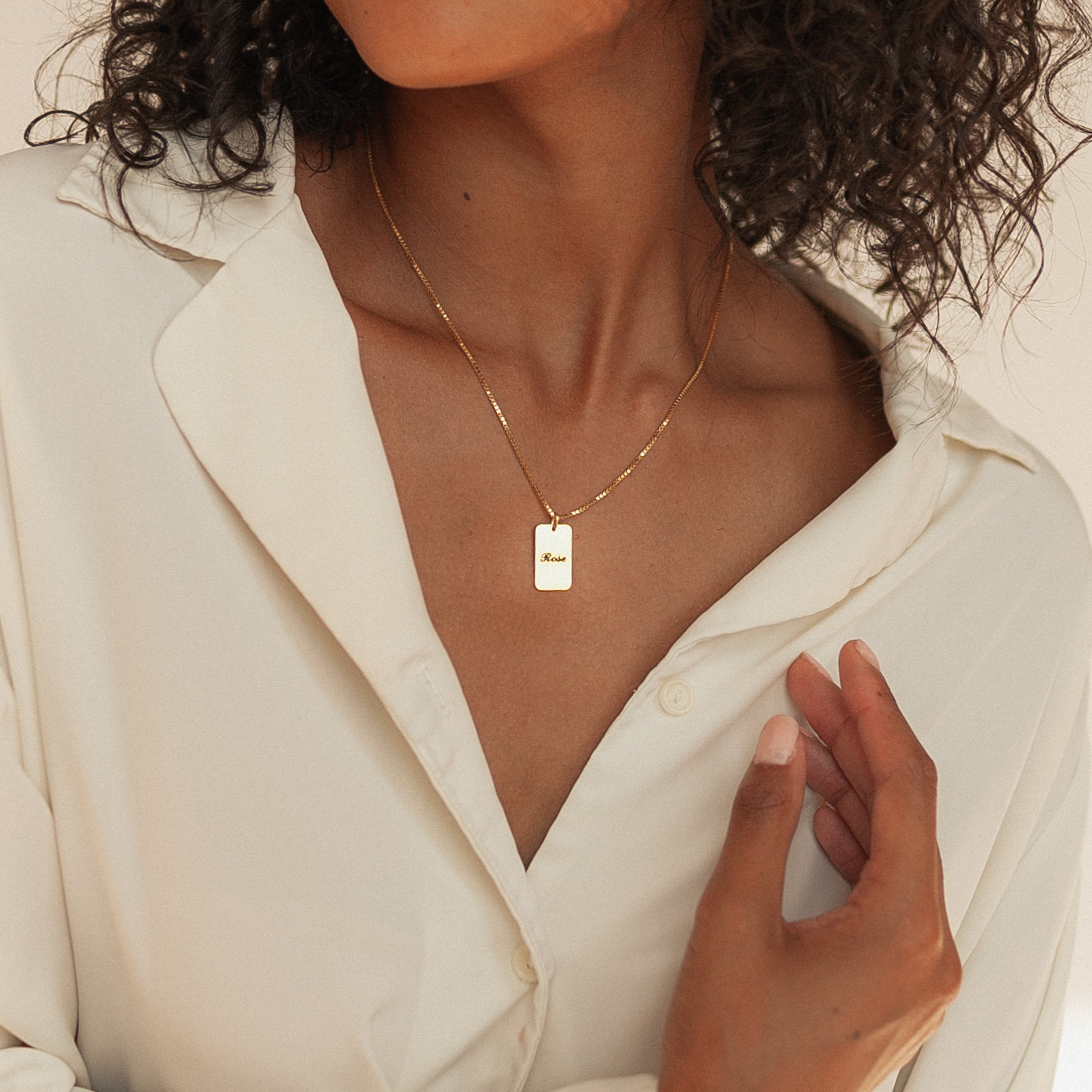 A woman in a cream blouse wears the Name Tag Necklace in Rose Gold, featuring a rectangular pendant, with her hand lifted near her collar.