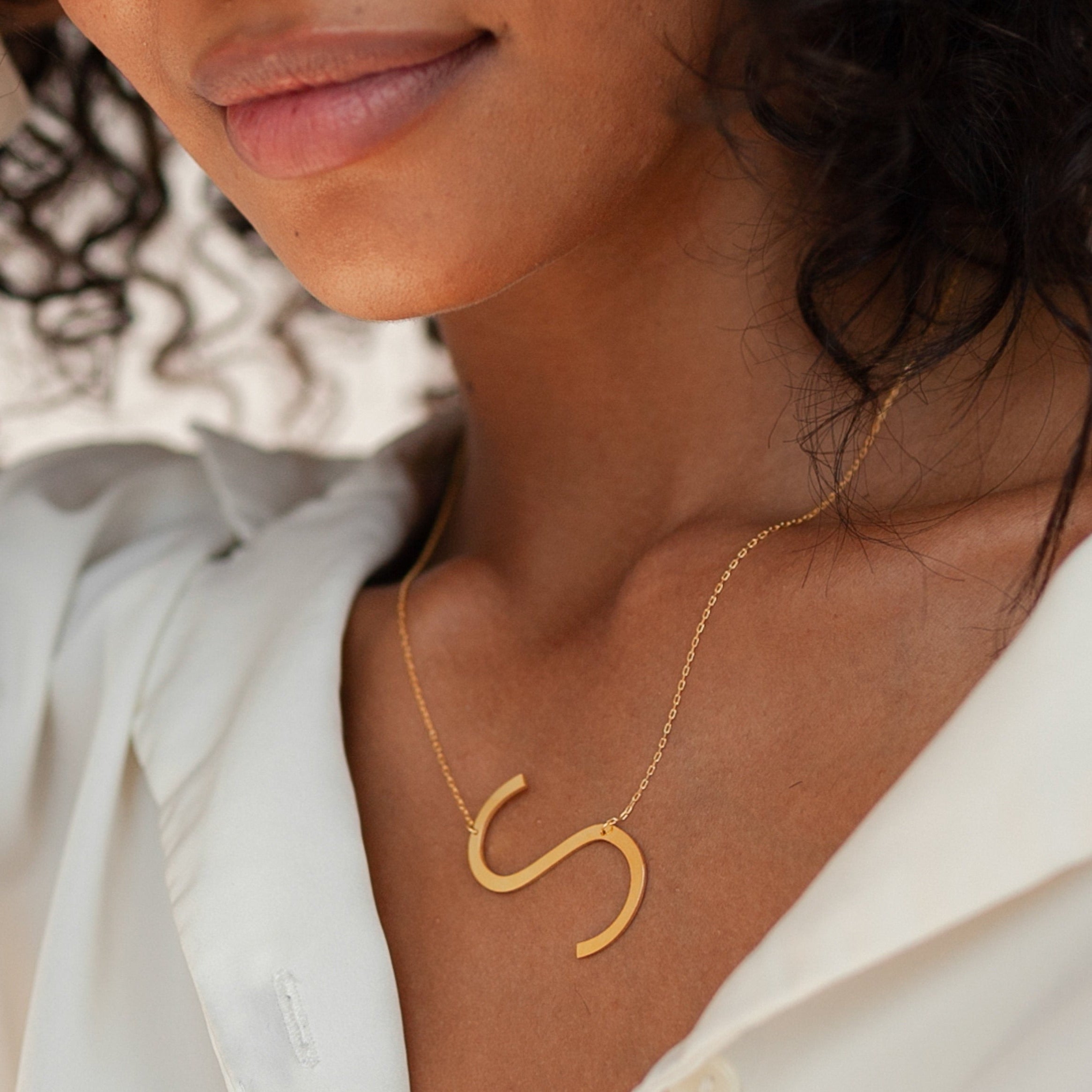 A woman wears a white blouse and the Large Sideways Initial Necklace featuring a bold gold S-shaped pendant—an effortless statement piece.