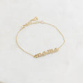 The Pave Mama Script Bracelet in gold, with "mama" spelled out in sparkling detail, rests on white marble—a stylish and elegant piece of jewelry perfect for celebrating motherhood.