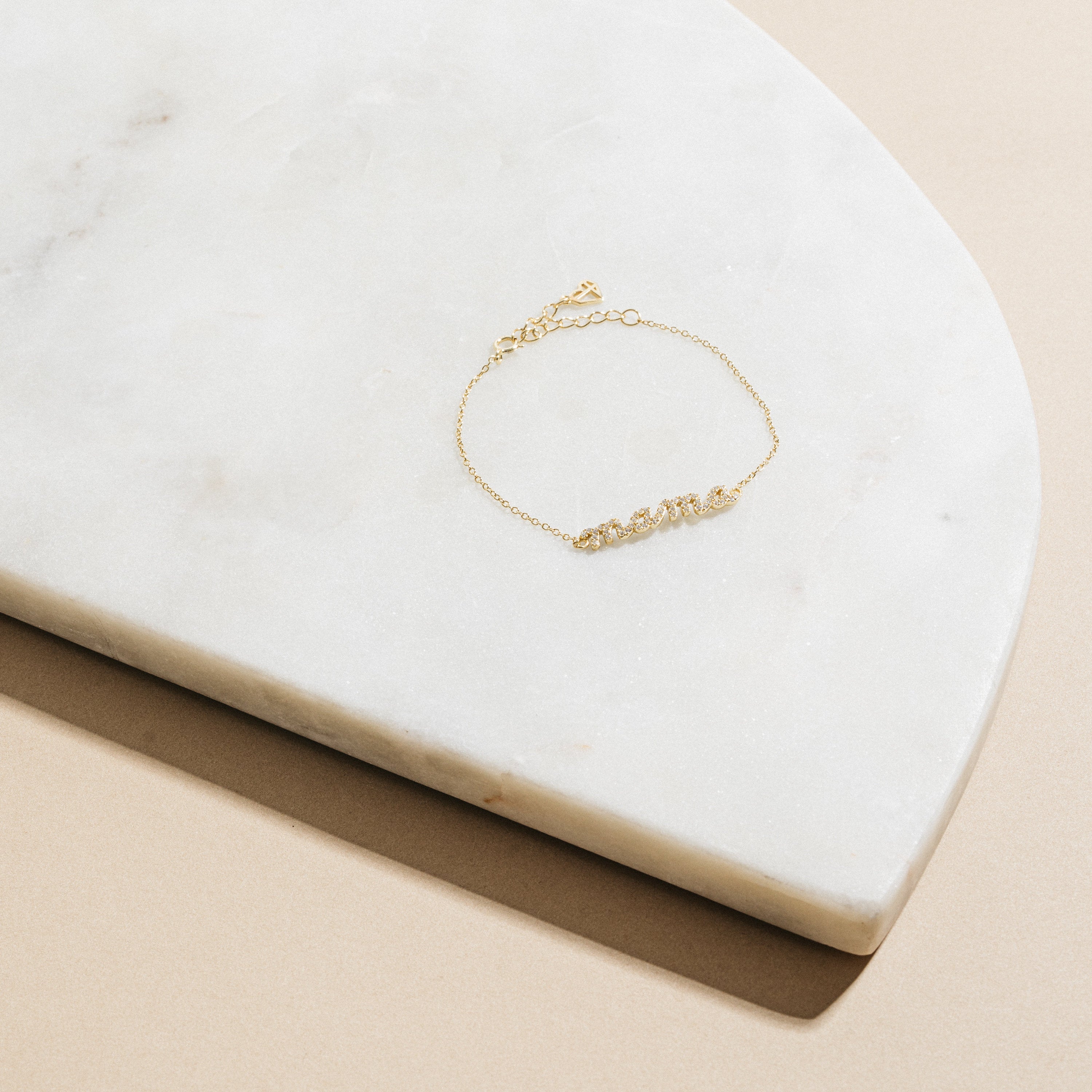 The Pave Mama Script Bracelet in gold, with "mama" spelled out in sparkling detail, rests on white marble—a stylish and elegant piece of jewelry perfect for celebrating motherhood.