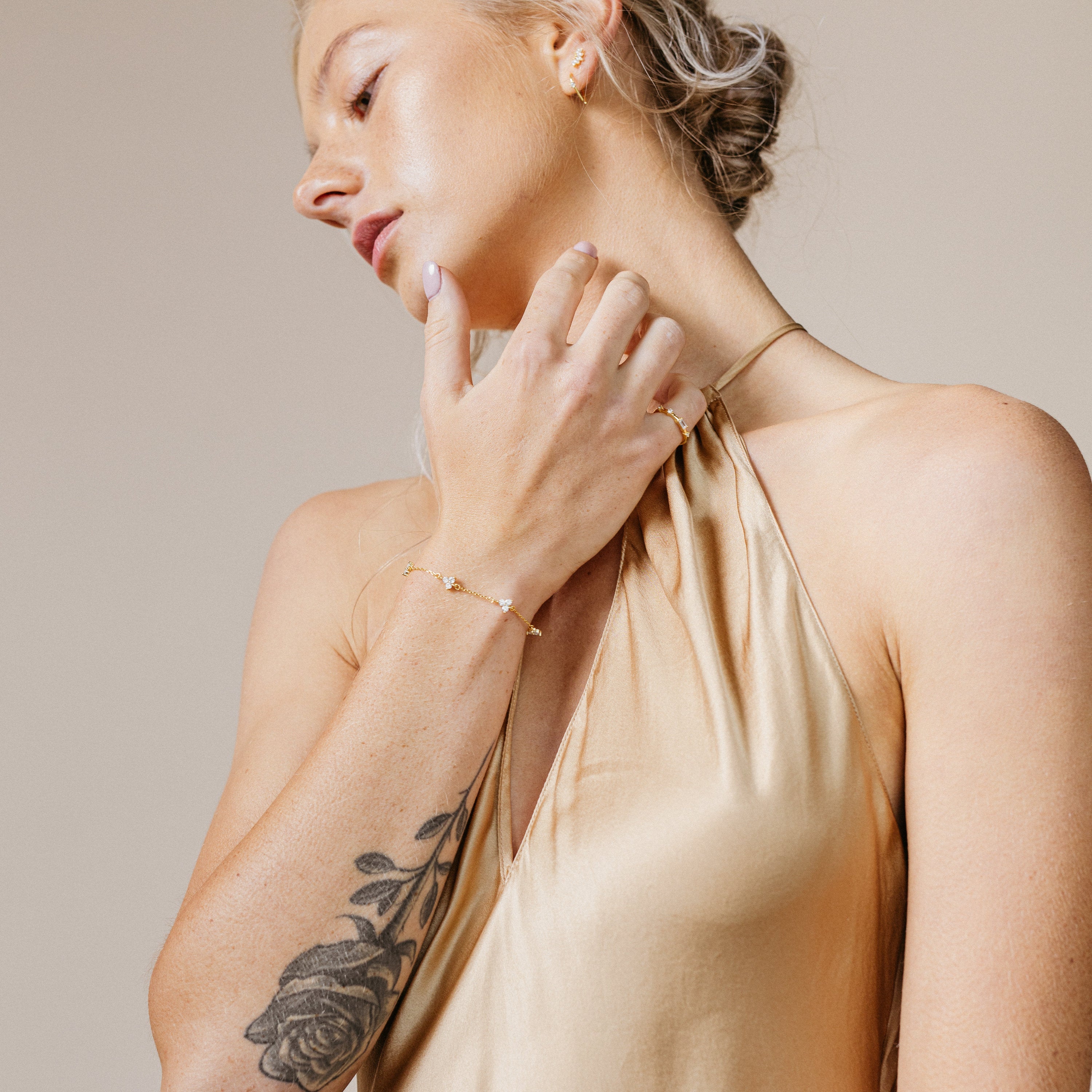 Woman in a gold halter dress touches her neck, showing a rose tattoo and subtle gold jewelry, with the Mini Lily Diamond Bracelet elegantly adorning her wrist.