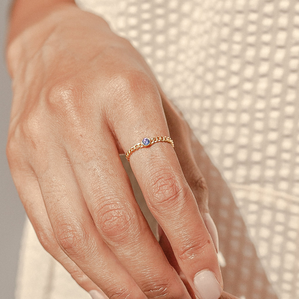 A hand models the Birthstone Curb Ring—a delicate gold curb chain ring set with a small purple gemstone—against a textured cream fabric background.