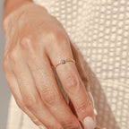 A hand models the Birthstone Curb Ring—a delicate gold curb chain ring set with a small purple gemstone—against a textured cream fabric background.