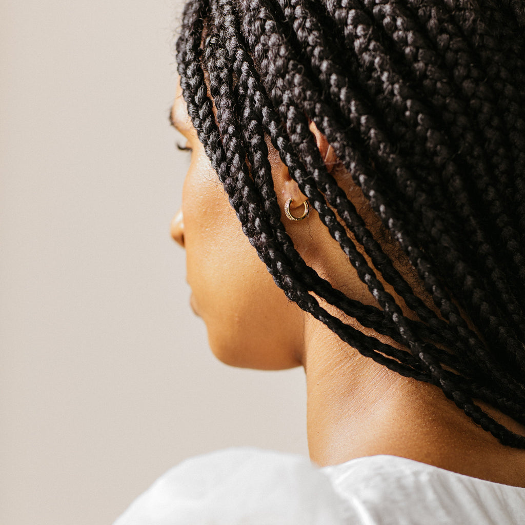 A woman with braided hair and gold hoop earrings, seen from behind in a white top, models the Coco Pave Huggies—perfect for an effortless everyday earring look.