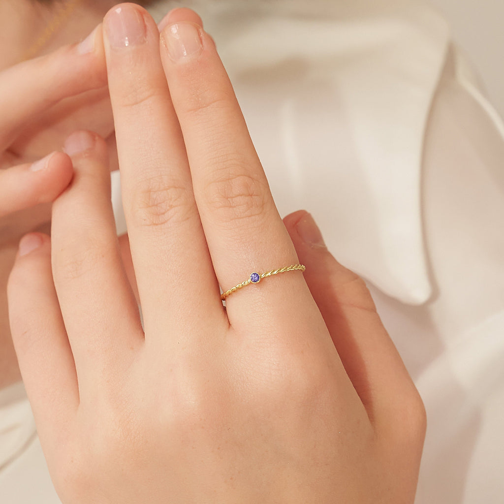 A hand wears the Twisted Birthstone Ring, a delicate gold design featuring a small blue gemstone—ideal for enhancing your ring stack.