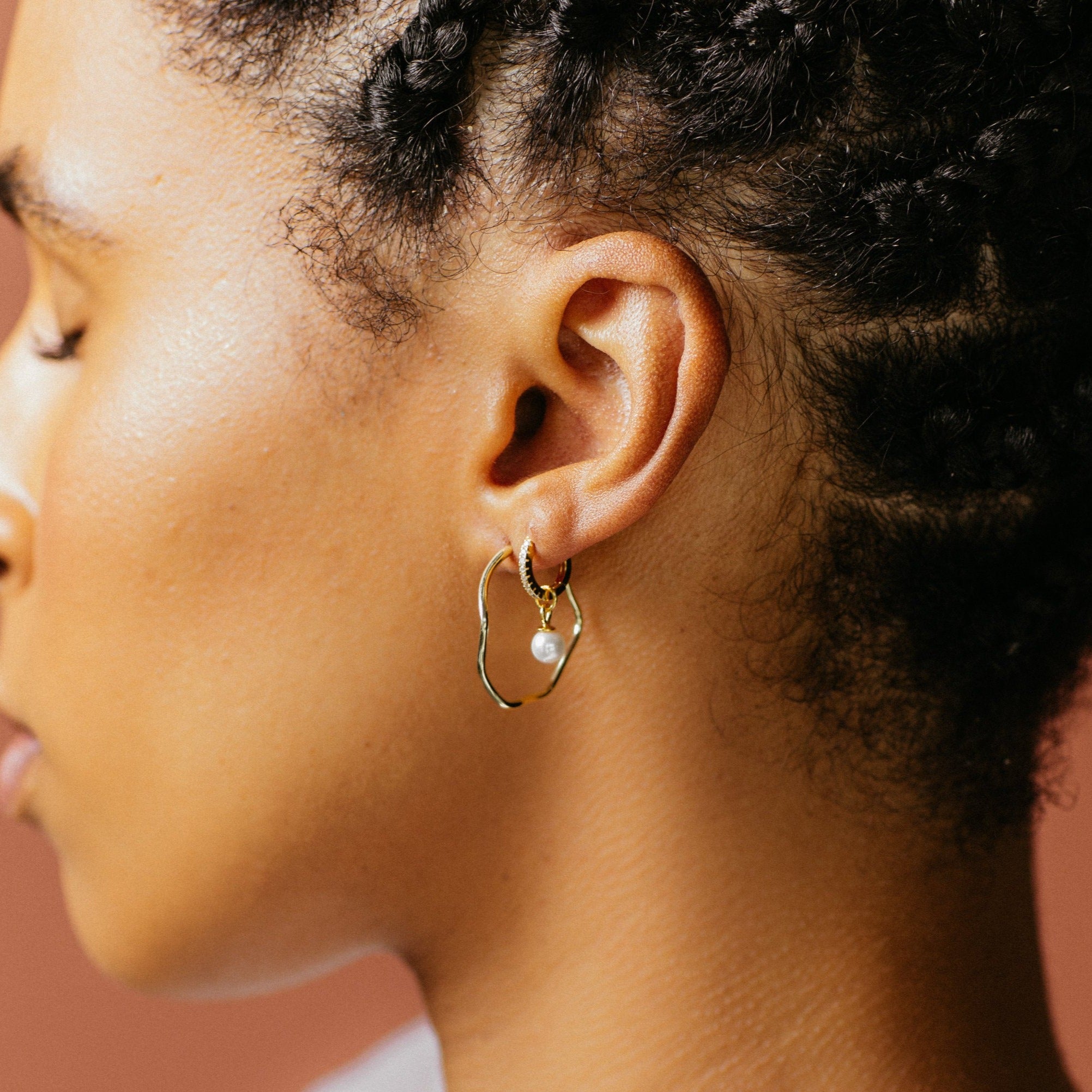 Close-up of a person wearing Mae Huggies—elegant gold hoops with pearls—highlighting braided hair and smooth skin.