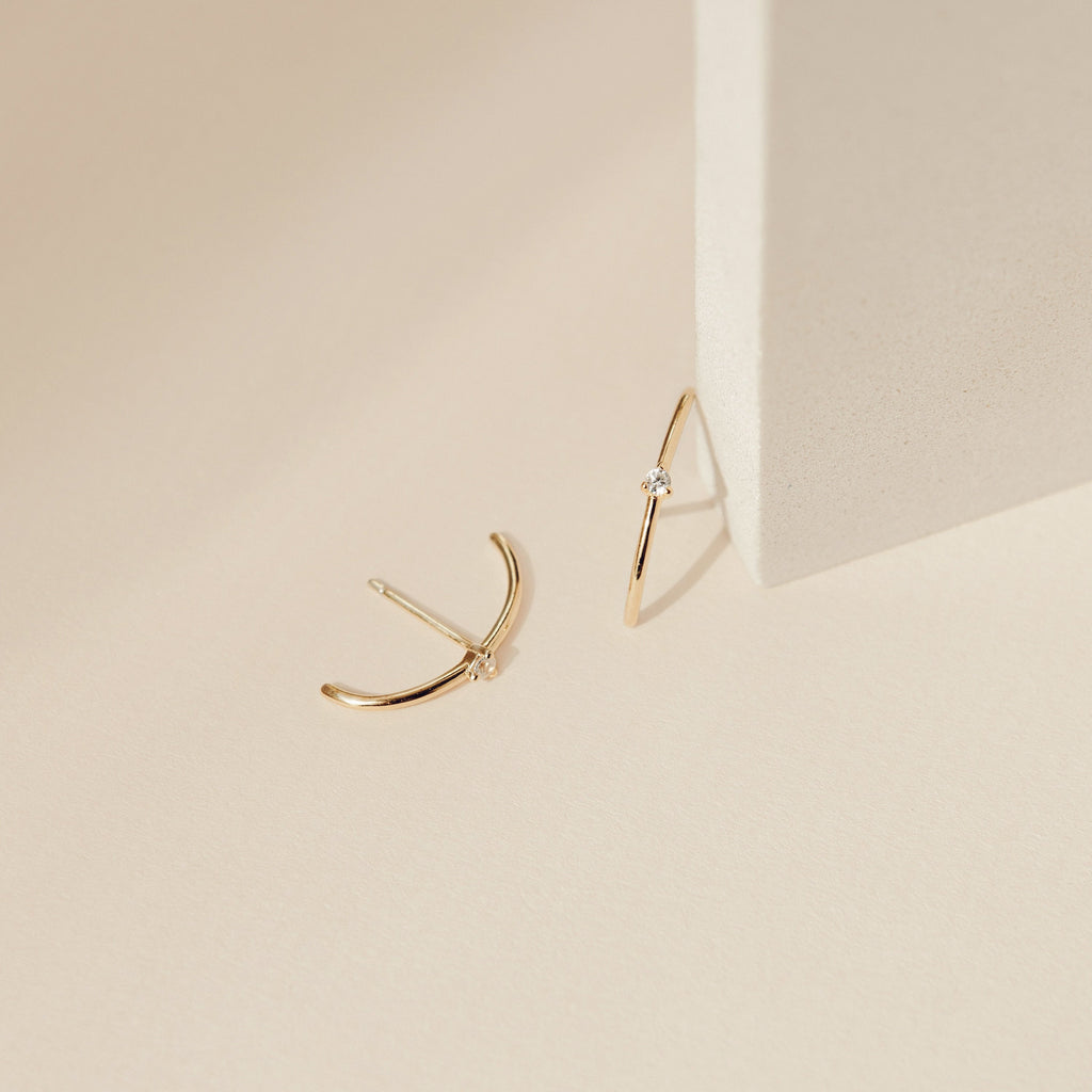 The Arc Studs, minimalist gold arcs with small diamonds, are shown on a light beige surface beside a block—ideal for fans of minimal ear jewelry.