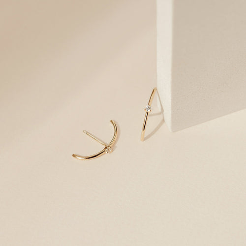 The Arc Studs, minimalist gold arcs with small diamonds, are shown on a light beige surface beside a block—ideal for fans of minimal ear jewelry.