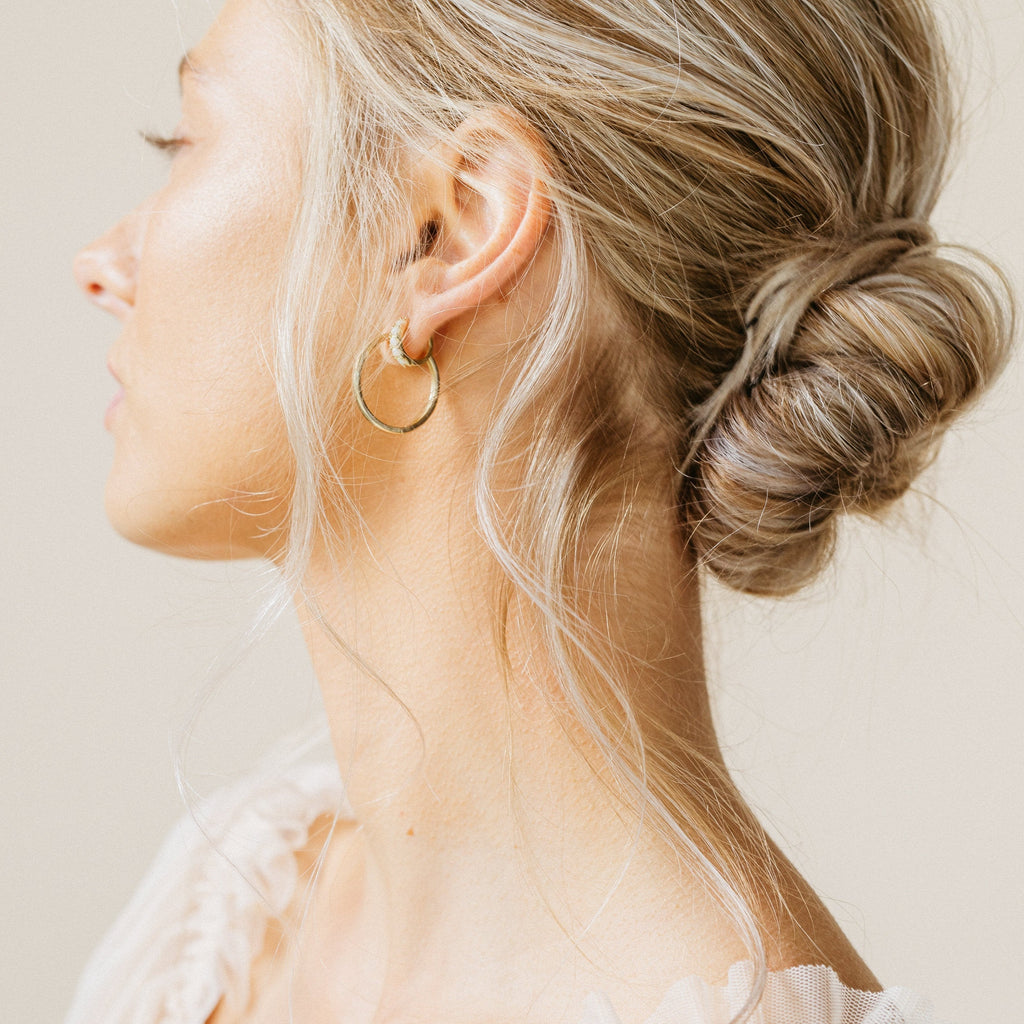 A woman with blonde hair in a low bun wears Opal Huggies earrings, facing sideways against a light background.