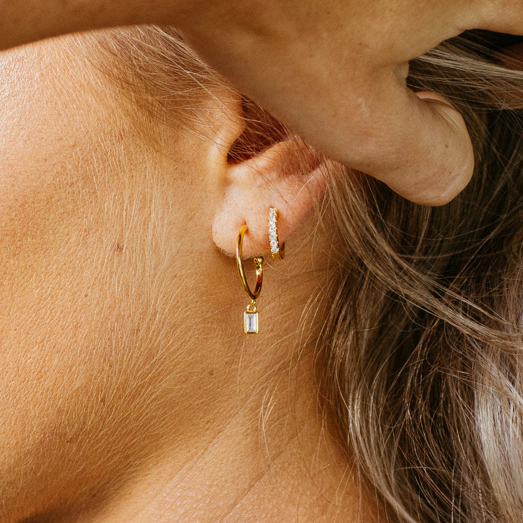 Close-up of a person’s ear adorned with two gold hoop earrings, including Coco Pave Huggies featuring a small rectangular pendant for an effortlessly chic everyday style.