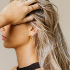 Woman with blonde hair wears gold earrings—Elle Huggies—and a matching ring, her hand gently covering part of her forehead in a graceful side profile.