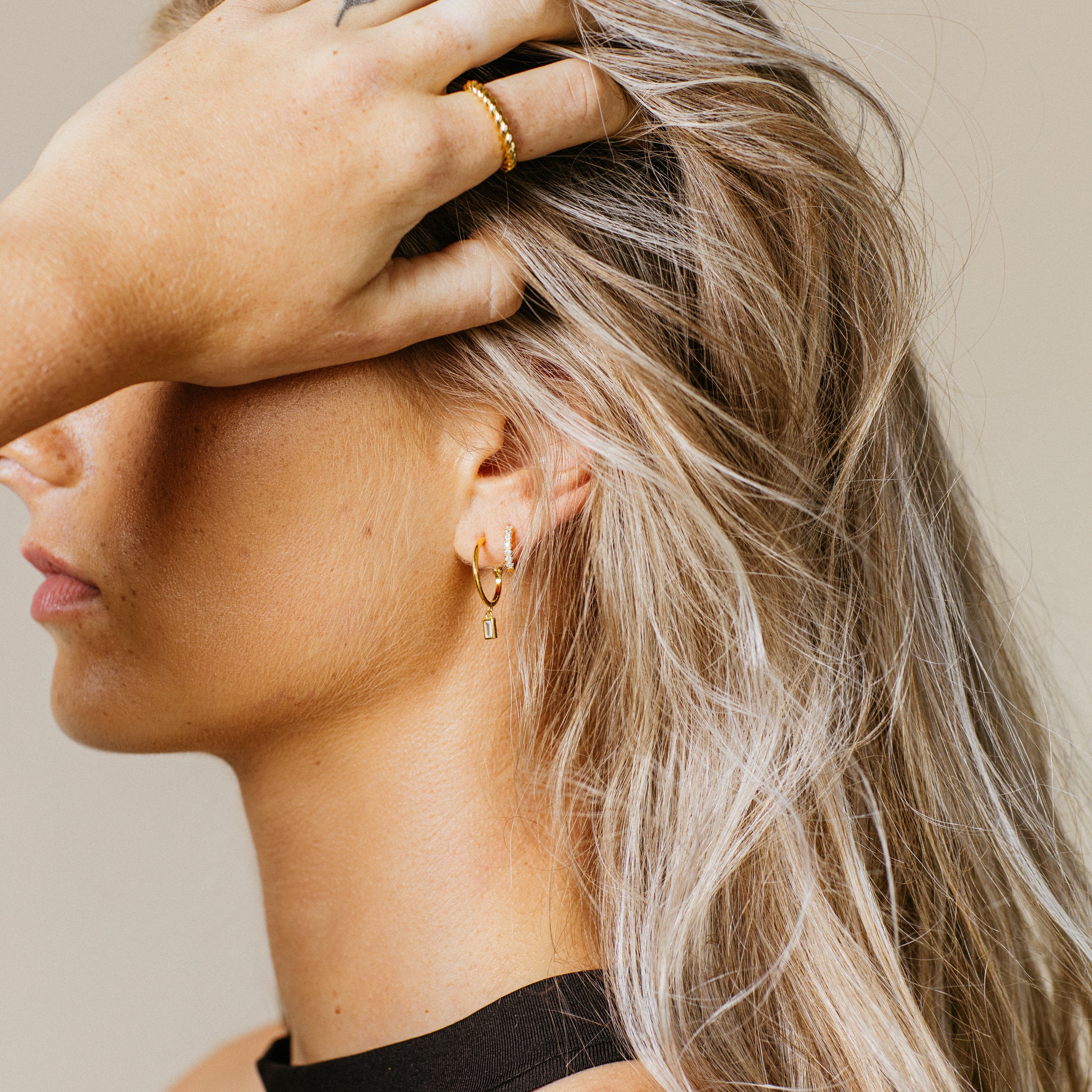 Woman with blonde hair wears gold earrings—Elle Huggies—and a matching ring, her hand gently covering part of her forehead in a graceful side profile.