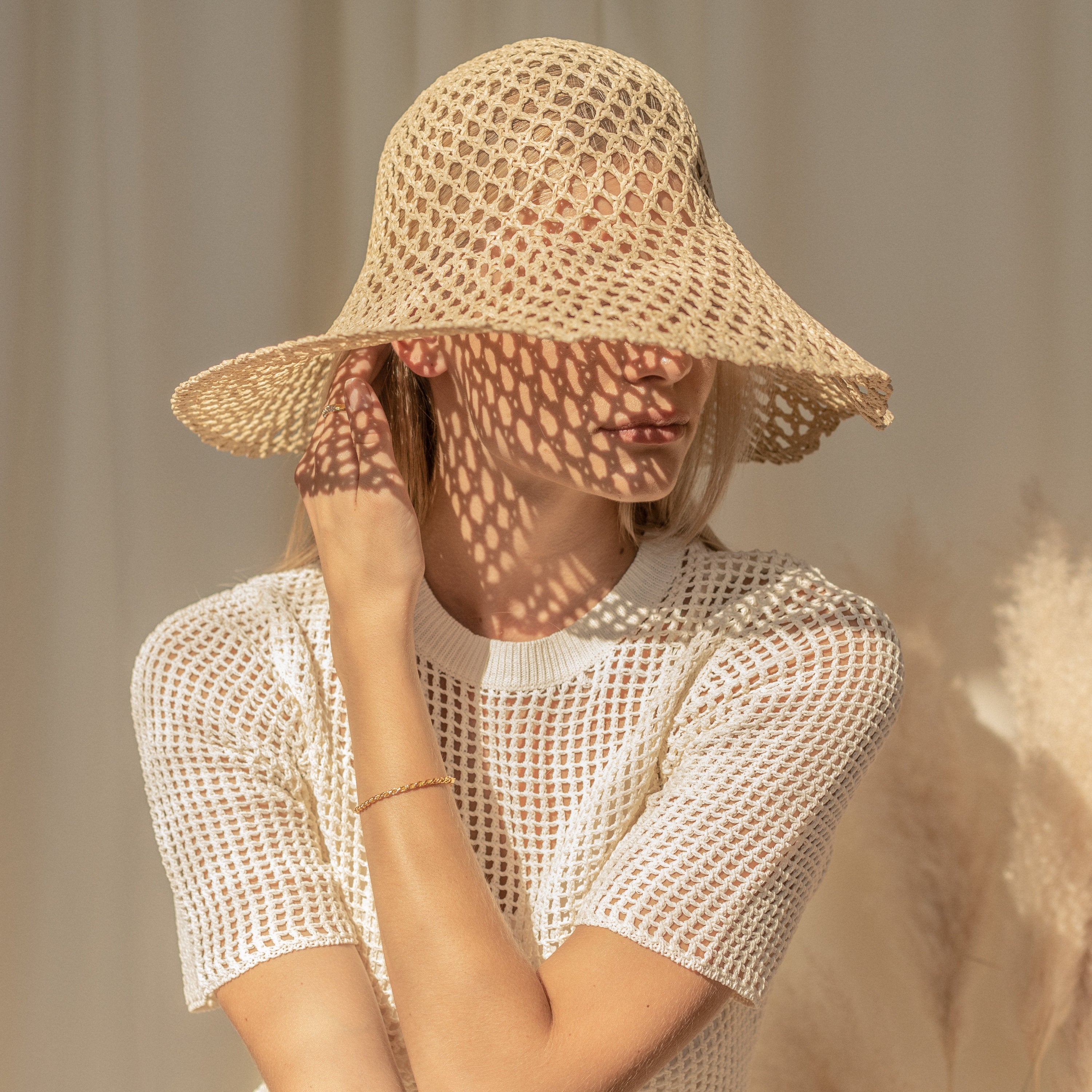 A woman in a white mesh top and woven sun hat, shadow patterns on her face, touches her ear while wearing the Rope Chain Bracelet, known for its elegant twisted design.