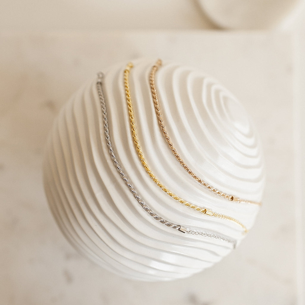 Three Rope Chain Bracelets—one silver, one gold, and one rose gold with a subtle twisted design—are draped over a white textured sphere.