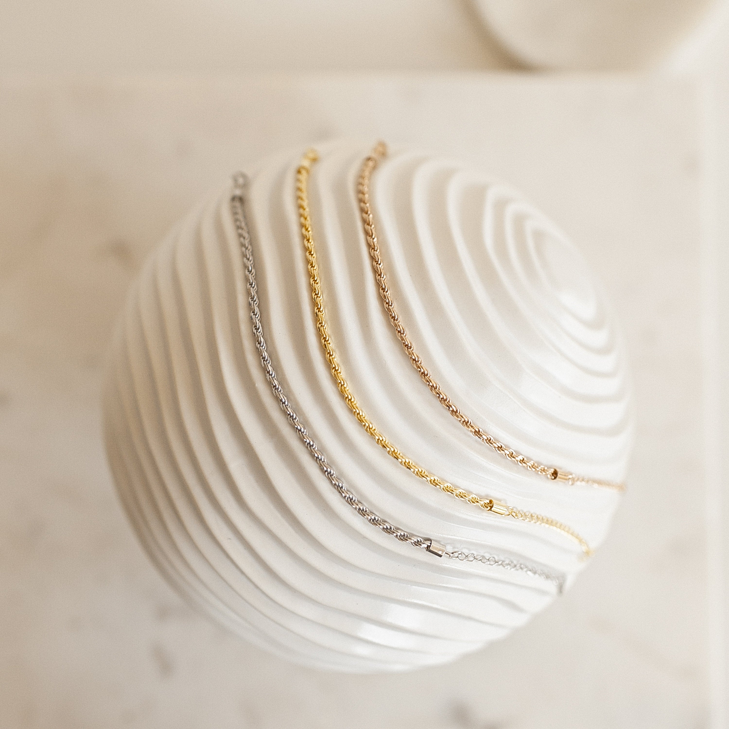 Three Rope Chain Bracelets—one silver, one gold, and one rose gold with a subtle twisted design—are draped over a white textured sphere.