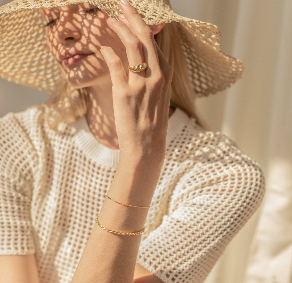 A woman, adorned in a woven hat and white knit top with gold jewelry—including the Box Chain Bracelet—sits in sunlight as delicate shadows fall across her face.