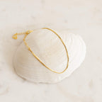 The Essential Bracelet Chains, featuring a delicate gold design with polished finishes, are displayed on a white seashell set against a light, neutral background.
