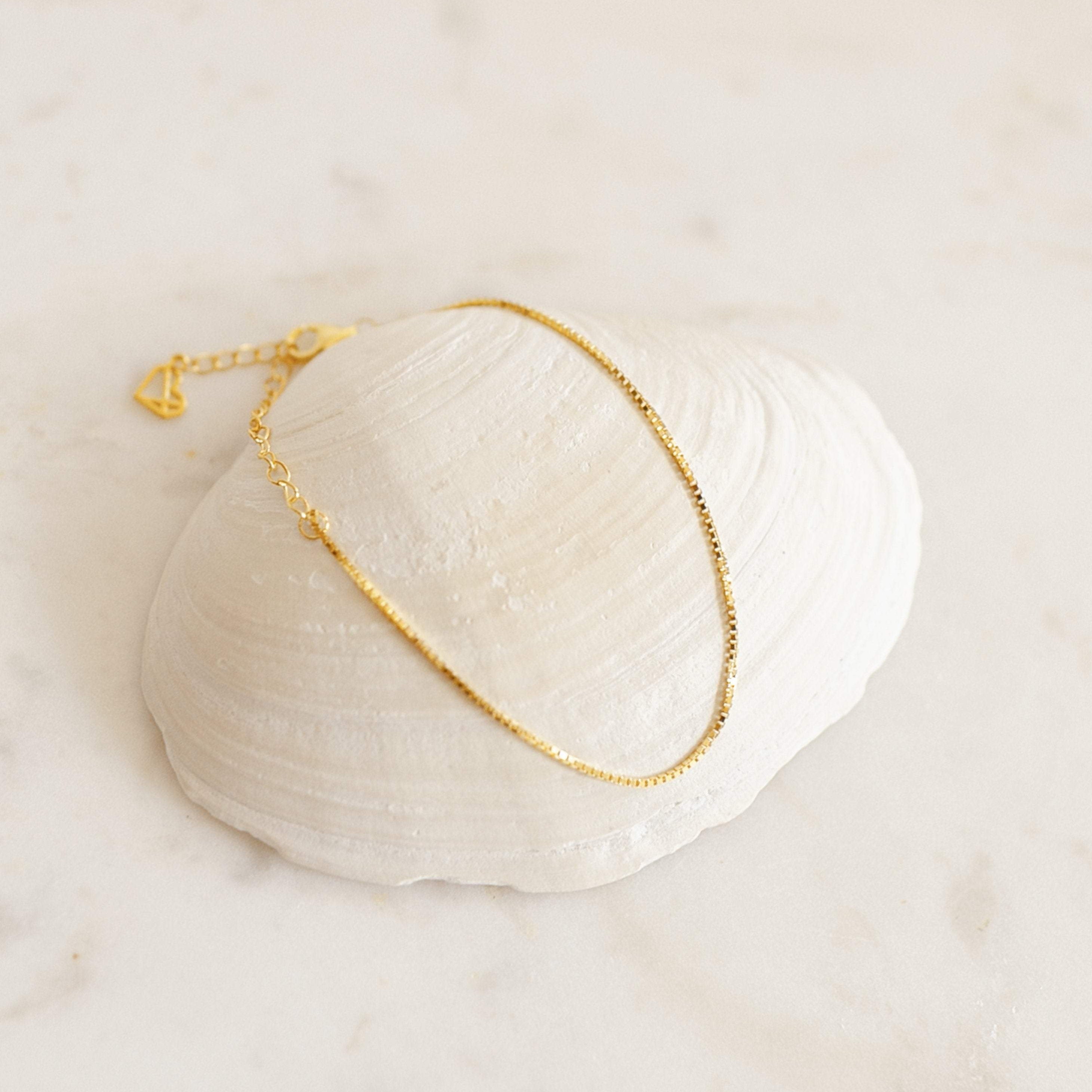 The Essential Bracelet Chains, featuring a delicate gold design with polished finishes, are displayed on a white seashell set against a light, neutral background.