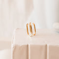 Nina Link Earrings, featuring sleek rectangular gold huggies, are displayed on a white textured surface with a soft, neutral background.