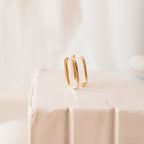 Nina Link Earrings, featuring sleek rectangular gold huggies, are displayed on a white textured surface with a soft, neutral background.