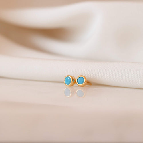 Ariel Turquoise Studs—dainty gold earrings with round turquoise stones—are elegantly displayed on white fabric, perfect for lovers of turquoise jewelry.