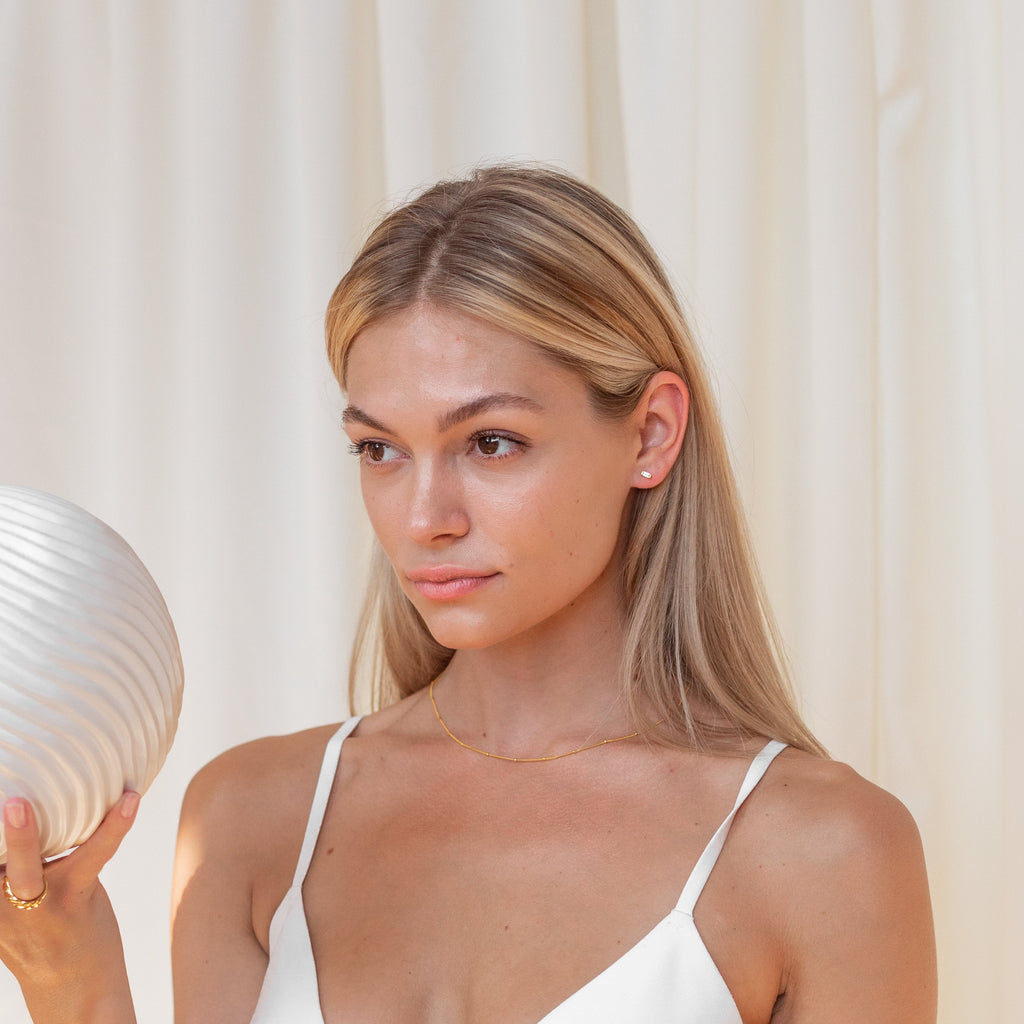A woman in a white top holds a textured white vase before cream-colored curtains, her look finished with Baguette Diamond Studs.