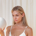 A woman in a white top holds a textured white vase before cream-colored curtains, her look finished with Baguette Diamond Studs.