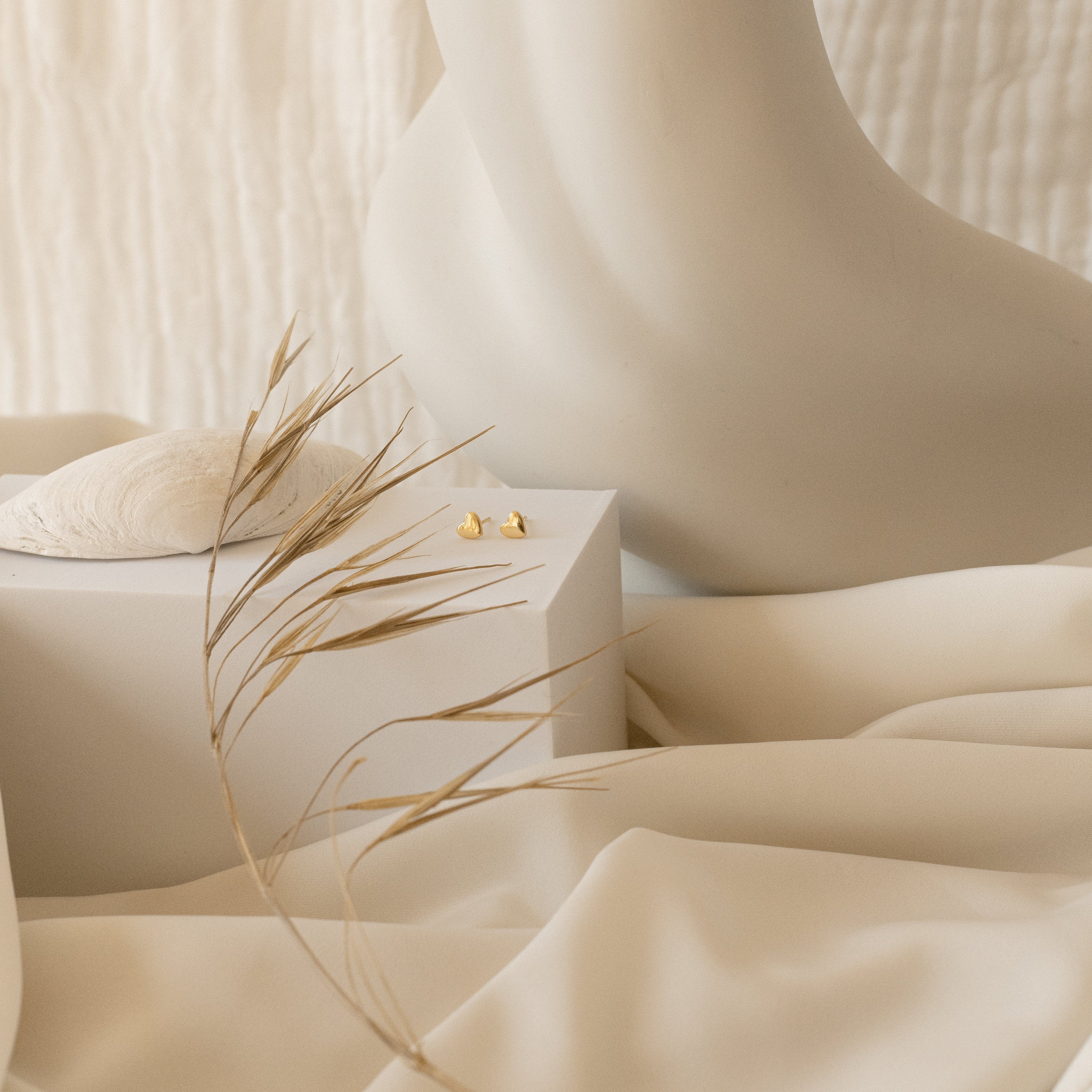 Minimalist scene featuring cream fabric, a sculpted figure, dried grass, a seashell, and the Classic Heart Studs on a box—an elegant backdrop to highlight these cute earrings.